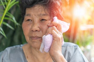 Elderly woman using a towel with ice pack on her face for pain relief, showing home healthcare,...