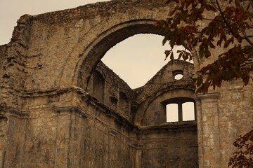 The weathered stone ruins of a historic castle or church with large arched openings against a pale sky, framed by dark autumnal tree branches in Kastav, Croatia.