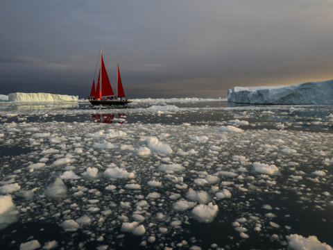 Red-sailed boat sailing through icy arctic waters surrounded by floating ice and large icebergs under cloudy sky.