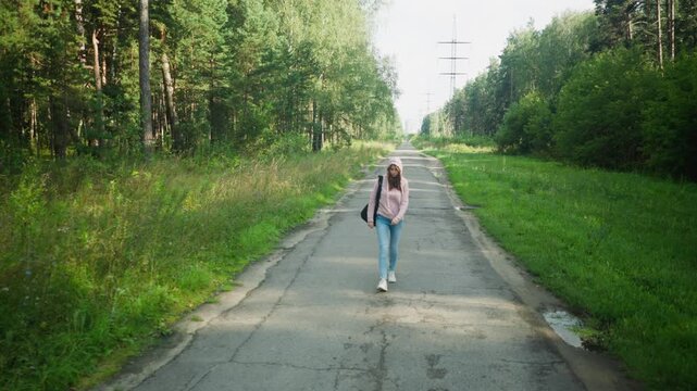 Young woman in pink hoodie and jeans walking along empty forest road, holding her hoodie rope thoughtfully, casting long shadow on pavement under morning sunlight, surrounded by trees and wild grass