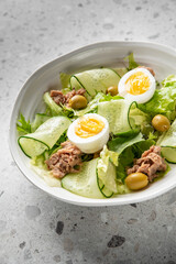 Fresh salad with tuna, cucumber ribbons, boiled eggs, olives, and lettuce in a white bowl on a gray surface. Healthy protein-rich meal, top view, vibrant food styling