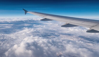 Obraz premium Aerial view showing an airplane wing soaring above clouds, with a mountainous landscape visible below against a blue sky backdrop