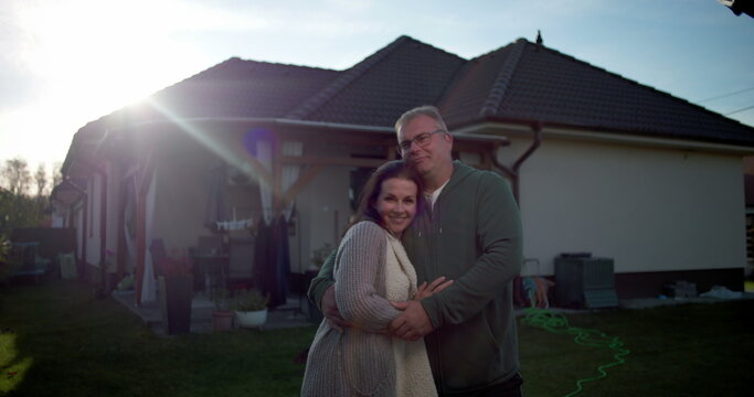 Couple embracing in backyard, standing near residential house, warm and affectionate moment in suburban home setting with patio, lawn, and greenery