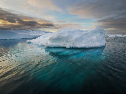 Massive iceberg in calm Arctic waters with deep blue submerged base under a pastel-colored sky.