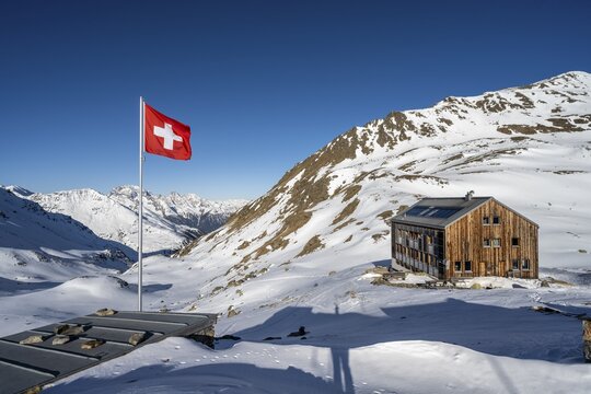 Swiss flag and mountain hut Keschh&uuml;tte in front of mountain landscape in winter, behind summit of the Berg&uuml;ner St&ouml;cke Tinzenhorn and Piz Ela, B&uuml;ndner Haute Route, Albula Alps, Rhaetian Alps, Grisons, Eastern Switzerland, Switzerland