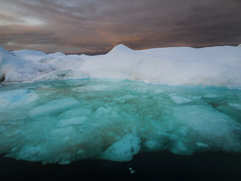 Arctic iceberg with deep turquoise submerged base in calm ocean under dramatic sunset sky with clouds.