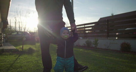 Father helping toddler walk on lawn near family home, supporting child’s movement and balance, fostering bonding and love, early childhood development outdoors