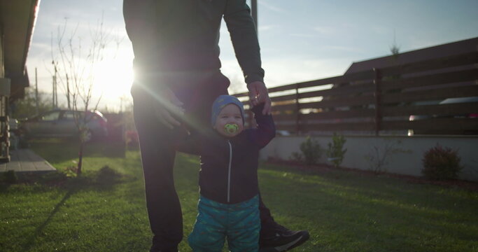 Father helping toddler walk on lawn near family home, supporting child’s movement and balance, fostering bonding and love, early childhood development outdoors - Powered by Adobe