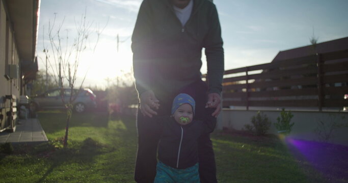 Father helping toddler walk on lawn near family home, supporting child’s movement and balance, fostering bonding and love, early childhood development outdoors