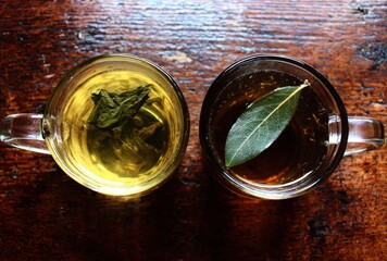 Two cups of tea sit side-by-side on a wooden table. One has green leaves in amber liquid and the other holds a large green leaf in darker tea