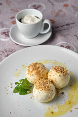 A delightful dessert, featuring round treats sprinkled with nuts, fresh mint, and a cup of coffee with whipped cream, all on a white plate.
