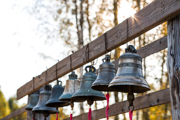 Obraz premium A row of antique bells is suspended on a wooden frame. The bells are of different sizes and are decorated with patterns. In the background, trees and sunlight can be seen.