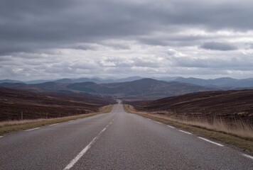 Naklejka premium Grey asphalt road extends toward distant hills under a cloudy sky, bordered by dry grasses and rolling terrain, perspective narrowing to horizon