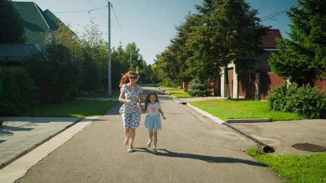 Young woman wearing floral dress running hand in hand with daughter in blue dress down quiet sunny residential street surrounded by green trees, suburban houses