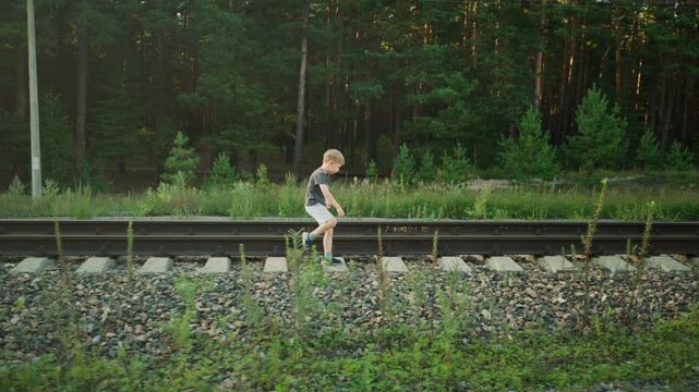 side view of little boy walking on rail sleeper along railway track in natural outdoor setting with dense forest in background and gravel stones on ground during peaceful afternoon scene