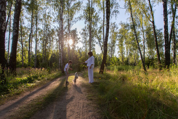 The family enjoys a daytime walk in a sunny forest with a dog on a dirty path surrounded by tall trees.