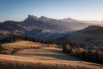 Golden sunlight illuminates a meadow landscape, with rugged mountains and distant snow-capped peaks in the background