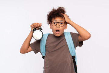 Scary socked African-American preteen boy male elementary primary pupil with bag holding round clock worried about the time harry up isolated over white background. Education back to school concept