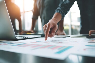 Three people examine colorful charts on a table with a laptop and phones. Sunlight creates highlights on the laptop. Depth of field blur