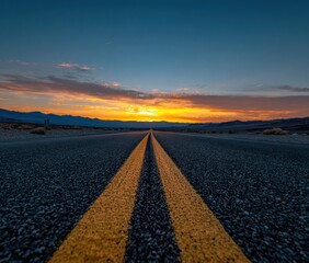 Asphalt road with a yellow line stretches toward a vibrant sunset over distant mountains, beneath a blue and orange sky