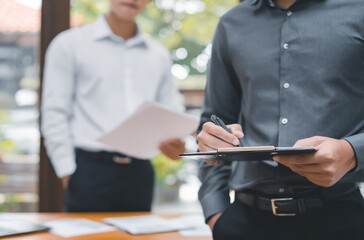 Two men in an office setting, one focused in the foreground signing a document on a clipboard, and the other man is partially visible in the background