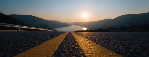 Low-angle view of a road with yellow lines leading to a lake surrounded by hills under a bright sunset. The road surface is visible in detail