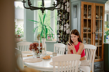 In a bright kitchen, a young woman sits at a white table, engrossed in her phone. A vase of flowers and snacks are nearby, adding warmth to the inviting space.