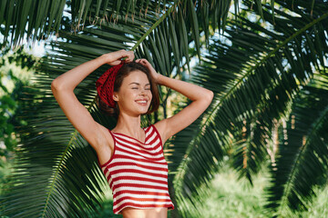 A cheerful woman enjoys a sunny outdoor tropical setting, wearing a red striped top and hat, posed among lush palm leaves with a bright, confident smile.