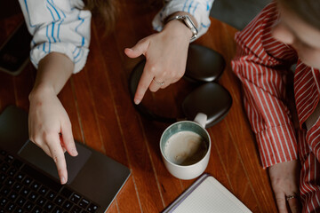 Two people engage in a discussion while pointing at a laptop. A coffee cup sits nearby on the wooden table, creating a cozy work atmosphere.