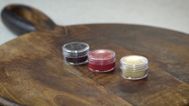 Three natural homemade lip balms in clear containers on brown background. DIY beauty products in red, brown, and transparent shades, organic skincare, handmade cosmetics, and eco-friendly style.