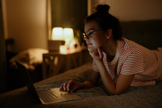 A young woman with glasses is lying on her stomach in bed, focused on her laptop in a dimly lit room. Soft light from a nearby lamp creates a cozy atmosphere. - Powered by Adobe