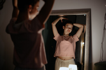 A young woman stands in front of a mirror, arms raised, as she admires her reflection in a softly lit room. The atmosphere is calm, highlighting her thoughtful expression during the evening.