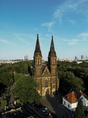 Naklejka premium Twin Gothic spires of the Vyšehrad basilica rising above the trees with city skyline in background.