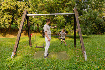 A father engages with his child at a playground, encouraging them while swinging. They share joyful laughter amidst greenery, highlighting the bond they have in this outdoor setting
