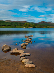 The calm, tranquil waters of a Loch Brora shoreline against rolling rugged hills and heather-clad moorlands of the Highlands in Northern Scotland. Bright blue sky.
