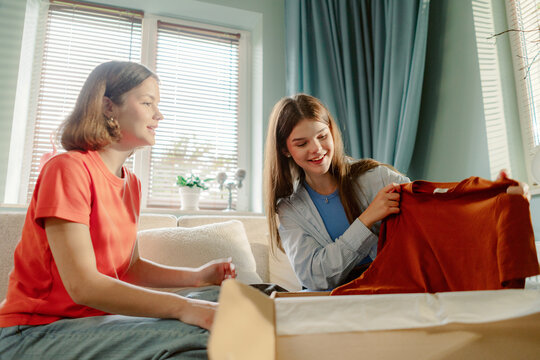 Two friends sit together in a cozy living room, excitedly unpacking new clothing. They share smiles and laughter, showcasing a relaxed atmosphere filled with friendship and joy.