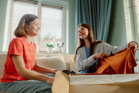 Two young women sit in a bright living room, joyfully unpacking a cardboard box filled with new clothes. They share smiles and laughter as they explore their latest fashion finds together.