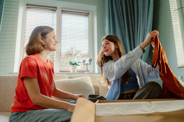 Two young women are seated in a cozy living room, unpacking clothes from a box. They are smiling and engaging in conversation, creating a warm and friendly atmosphere.