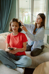 Two friends relax in a bright living room. One is sitting cross-legged using a phone while the other styles their hair with a curling tool. The atmosphere is warm and casual.