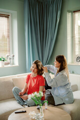 Two friends relax in a bright living room while one styles the other's hair. They sit comfortably on a sofa, surrounded by natural light and greenery, enjoying their time together.