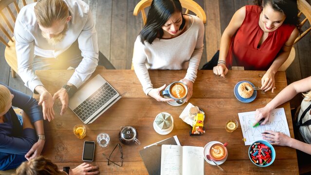 Diverse group having a meeting over coffee. Top view of people with laptops and notes. Collaborative discussion at cafe. Business meeting at coffee shop. Diverse men and women discuss at cafe - Powered by Adobe