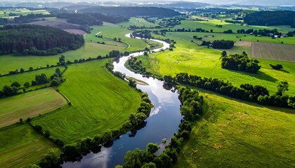 Obraz premium Aerial view reveals a meandering river flowing through vibrant green fields and forests, under a partly cloudy sky