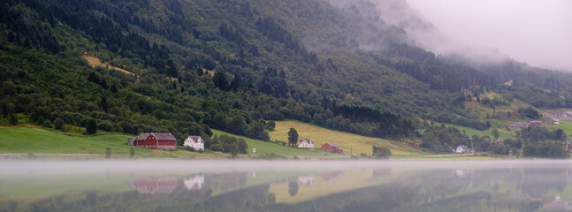 Calm waters and mist envelop Bergheimsvatnet, revealing beautiful farmhouses nestled against lush green hills. A perfect capture of Norways serene natural beauty at dawn.