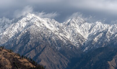 Obraz premium Majestic snow-capped mountains pierce the cloudy sky, contrasting with brown slopes and evergreen trees below. Serene and dramatic landscape captured