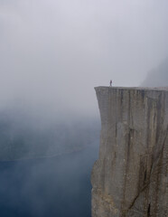A lone hiker stands at the edge of Preikestolen Cliff, surrounded by swirling mist and stunning...