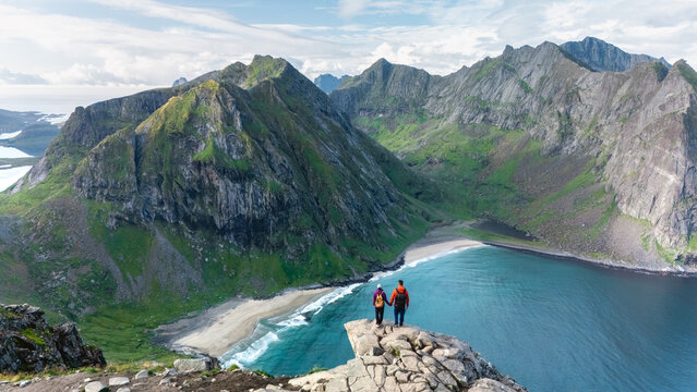 Hikers stand on a rocky ledge, gazing at the breathtaking landscape of Ryten Kvalvika Beach. The turquoise waters and rugged mountains create an unforgettable backdrop on this scenic trail in Norway.