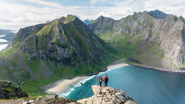 Two hikers admire the stunning landscape from Ryten peak, gazing at the turquoise waters of Kvalvika Beach. This unforgettable moment captures the beauty of Lofotens rugged terrain and lush greenery.