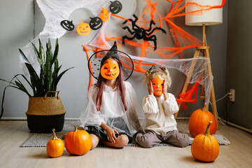 Two joyful children in Halloween costumes sit with pumpkins in a festive, decorated room. The image captures the fun and spooky atmosphere of a Halloween party indoors.