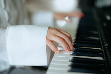 Obraz premium A person in a white shirt gracefully plays the piano, their fingers moving over the black and white keys. The bright room adds a warm atmosphere to the musical activity.