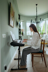 A woman sits on a chair, playing a keyboard in an elegant room filled with natural light. The space has stylish decor, featuring a chandelier and plants, reflecting a serene atmosphere.